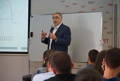 A lecturer in a suit presenting a graph to an audience at ISET, using a microphone and whiteboard for explanation