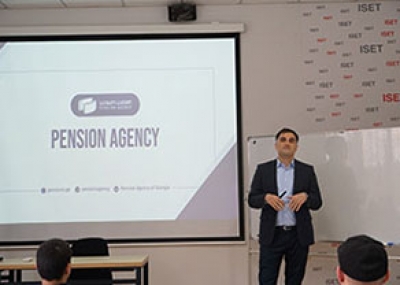 A speaker giving a presentation on the Pension Agency at ISET, with a projection screen displaying the topic and an audience listening.